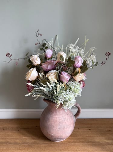 Customer's Faux Arrangement - Blush Rose with varied foliage displayed in a rustic clay pitcher on a wooden surface.