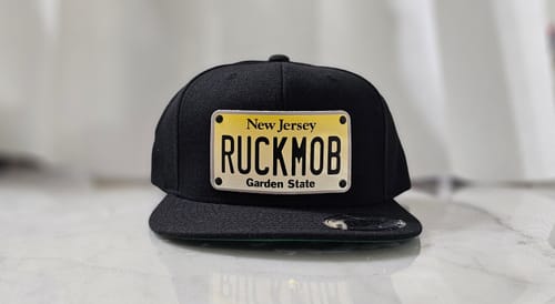 Customer's black New Jersey Plate Hat with a custom 'RUCKMOB' license plate patch on a marble surface.