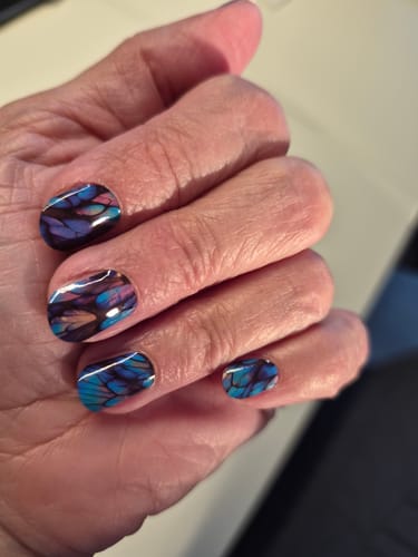 Hand einer Kundin, die die 'Blue Angel Wings'-Nagelfolien mit einem glänzenden, blau-violetten Design trägt.