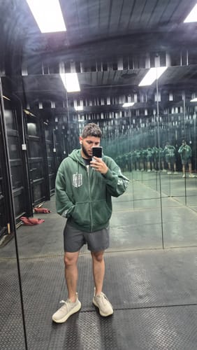 Customer takes a mirror selfie wearing the green AOT© x YLA Zip-Up Hoodie, showing its relaxed fit in a gym.