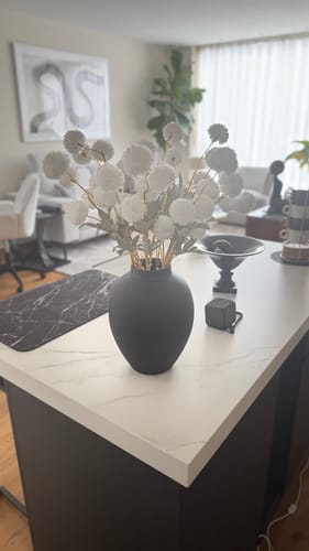 Recensent toont de witte Zijden Kiku Bloemen in een zwarte vaas op een keukeneiland.