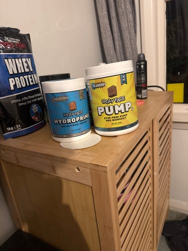 Reviewer's tub of UGLYFACE HYDROPRIME on a wooden cabinet with other workout supplements.