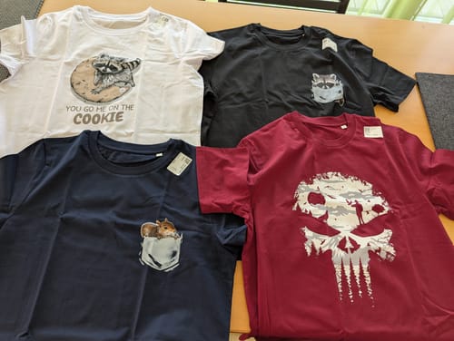 Kunde zeigt eine Auswahl von vier gekauften T-Shirts, darunter das schwarze „Frecher Waschbär“ Bio-Herrenshirt.