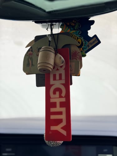 Reviewer's red 80Eighty air freshener hanging from a car mirror with other air fresheners.