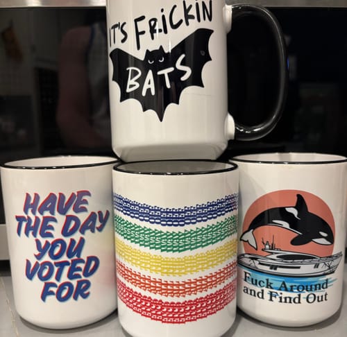 Reviewer's collection of four different Surprise Mugs, including designs for 'It's Frickin Bats' and 'Fuck Around and Find Out'.