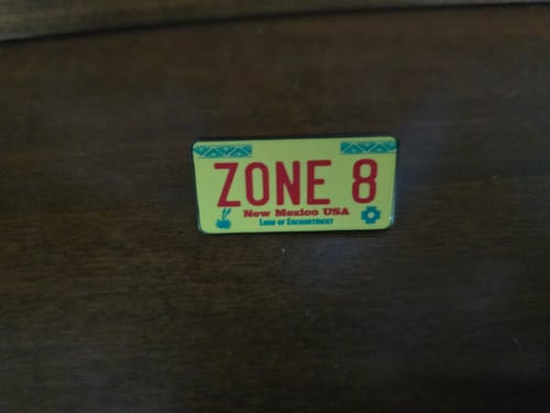 Customer's yellow New Mexico license plate enamel pin, customized with 'ZONE 8' in red text, resting on a dark wood surface.