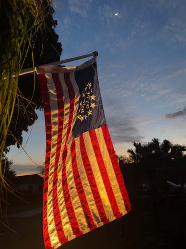 A customer's Betsy Ross Flag illuminated while hanging from a pole at dusk, with a crescent moon in the sky.