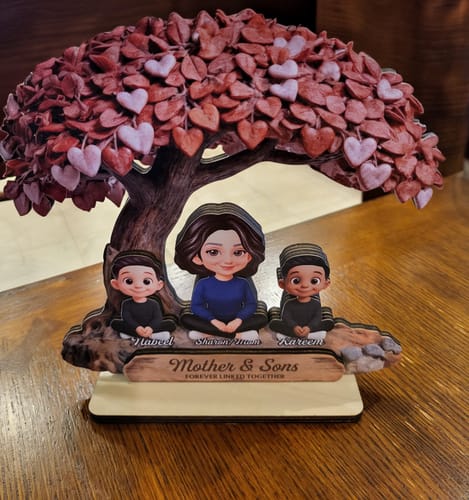 Customer's personalized wooden plaque with a mother and two sons under a heart tree, displayed on a wooden table.