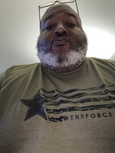 Close-up of a reviewer wearing the olive green Rugged O.G Tee, showing the fit and the black graphic on the chest.