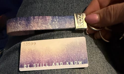 Reviewer's hand holding the sparkly 'Be Still and Know' bracelet next to its matching collector card.