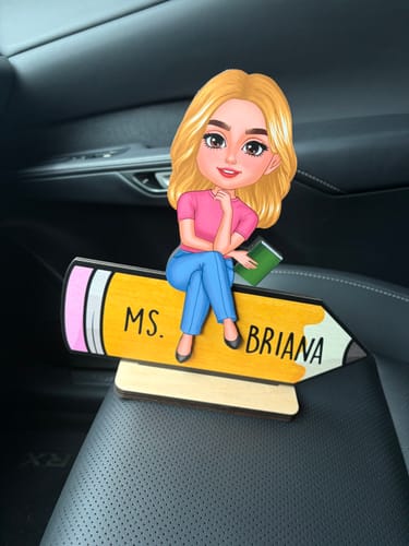 Customer's personalized Doll Teacher plaque with "MS. BRIANA" on a pencil design, shown sitting on a car seat.