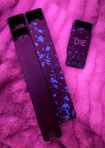 Reviewer's customized 2-Pack Bundle of black pill cases with a blue splatter design, shown on a pink fuzzy surface.