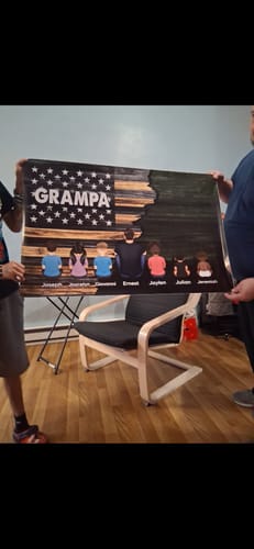 Two people holding a large personalized poster with 'GRAMPA' written over a flag design, showing an adult and children from behind.