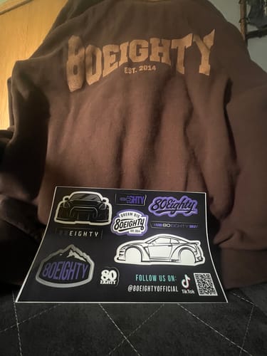 Reviewer showing the back of their brown 80Eighty Rooted Hoodie with a sheet of car-themed stickers in front.