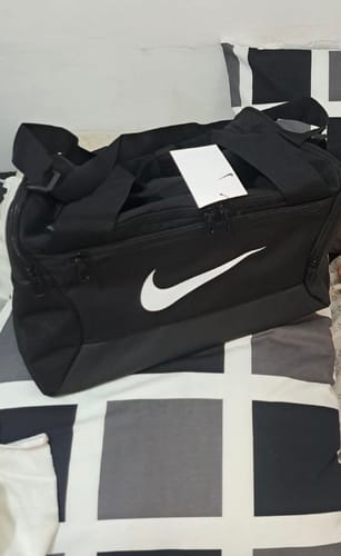 Reviewer's new black Nike Brasilia Duffel Bag with a white logo and tag, sitting on a bed with a patterned cover.