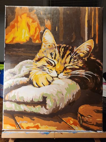 Reviewer's completed Cozy Dreams painting showing a tabby cat sleeping on a blanket by a fireplace.