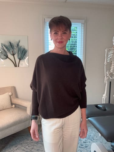 Reviewer standing in a room wearing the espresso brown Asymmetric Draped sweater with white pants.