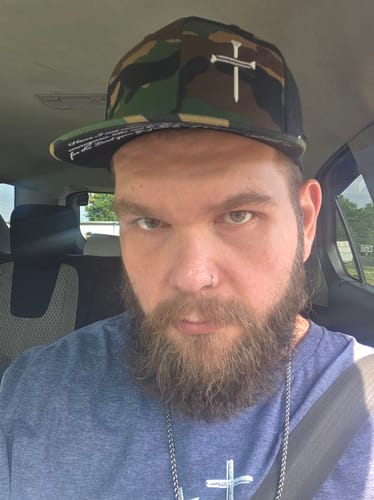 Reviewer in a car wearing the camouflage 3 Nail Cross Hat, showing the white cross and the prayer print under the brim.