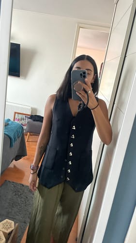 Una clienta se toma una foto en el espejo usando el chaleco VEST MANHATTAN negro sobre un pantalón verde.