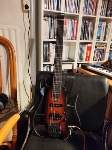 Ein Rezensent zeigt die Donner HUSH-X E-Gitarre mit Sunburst-Finish, die auf einem Stuhl steht.