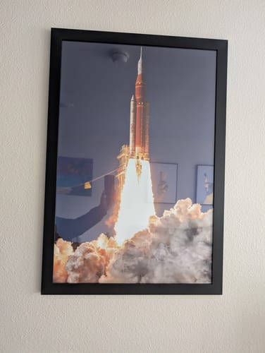 Reviewer's 'For All Humanity' print in a black frame, showing a rocket launch, hanging on a textured wall.