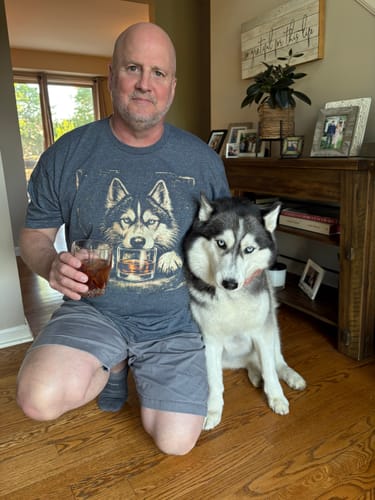 Reviewer wearing the grey Whiskey Husky t-shirt, kneeling next to a real husky and holding a drink.