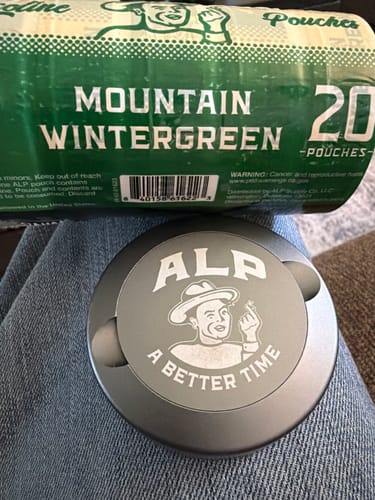 Customer's green roll of Mountain Wintergreen Nicotine Pouches and a grey ALP tin resting on their jeans.