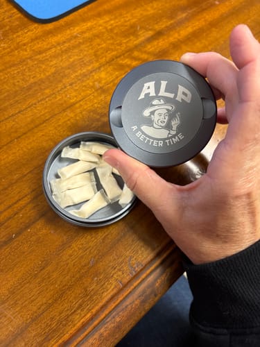 Customer's hand holding the lid of an open tin of ALP Tropical Fruit Nicotine Pouches on a wooden table.