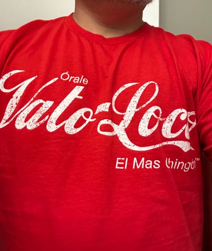 A reviewer shows a close-up of the red Vato Loco Art De La Calle™ Tee being worn, highlighting the white script logo.