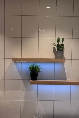 Kundens to stilige hyller med LED-lys og planter montert på en flislagt baderomsvegg.