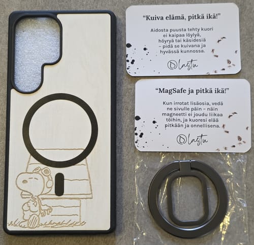 Arvostelijan puinen WOIMA MagSafe -kuori, johon on kaiverrettu Ressu-hahmo, sormuspidikkeen vieressä.