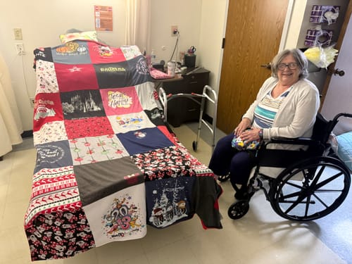Customer's photo of a smiling person in a wheelchair next to their Project Repat T-Shirt Quilt on a bed.