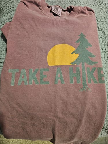 Reviewer's purple Take A Hike T-shirt laid out, showing the dull graphic of a sun and tree.
