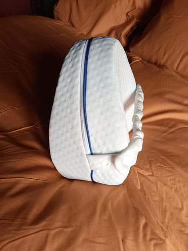 Une cliente montre son coussin orthopédique blanc pour genoux, posé sur un lit.
