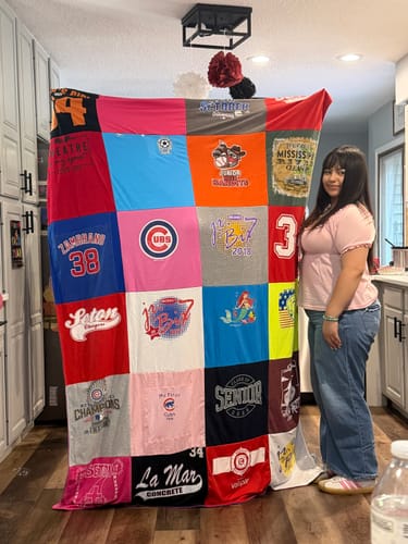 Reviewer standing next to her large, colorful Project Repat T-Shirt Quilt held up for display in a kitchen.