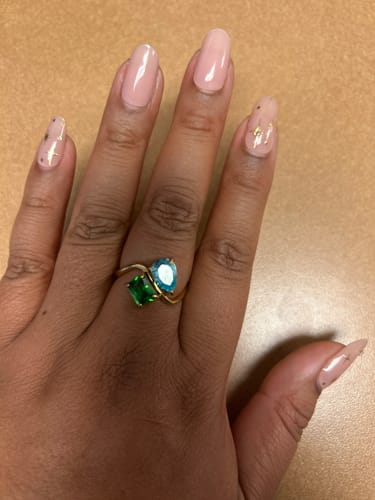 A customer's hand wearing the Navae Birthstone Ring, with a green radiant-cut stone and a light blue pear-cut stone.