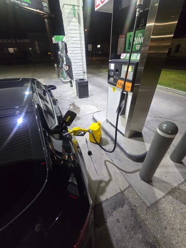 Reviewer's yellow 5 Gallon Jug with its spout inserted into a black car's fuel tank at a gas station.