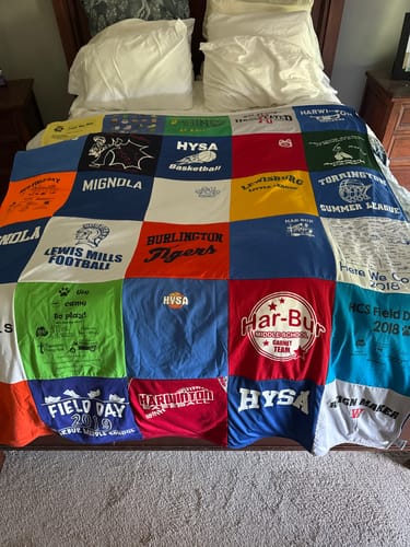 Customer's colorful Project Repat T-Shirt Quilt with various school and team logos spread on a bed.