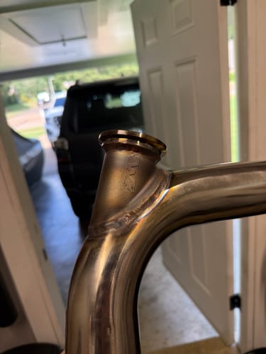Close-up of a customer's High Flow Wastegate Merge welded onto a shiny, curved steel pipe in a garage.