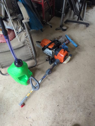 Reviewer's green 2.5 Gallon Jug on a concrete floor next to a string trimmer and a chainsaw, showing its use for two-stroke engines.