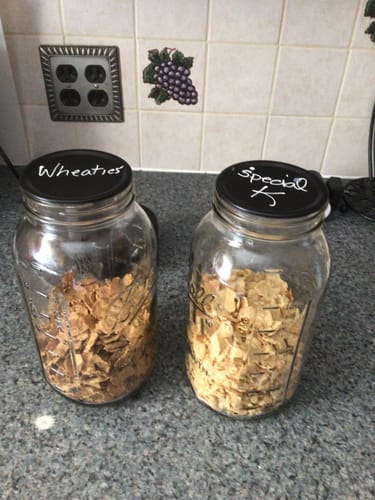 Customer's two glass jars of cereal with black lids labeled 'Wheaties' and 'Special K' using a white liquid chalk pen.