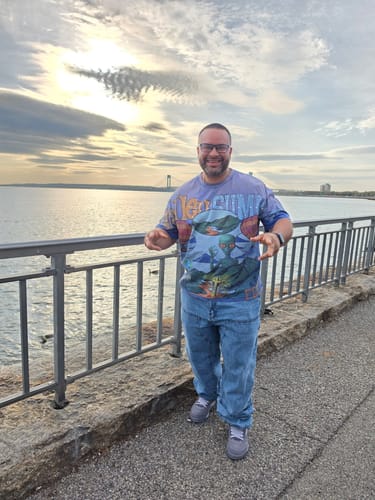 Reviewer standing outdoors by the water, wearing the colorful Hellstar Studios Records Tee with jeans.