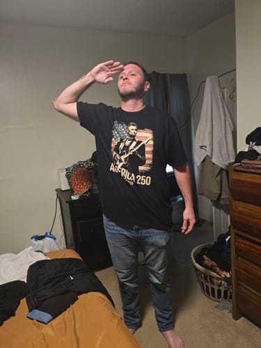 Reviewer saluting while wearing the black Freedom Rock 250 T-Shirt in a bedroom.