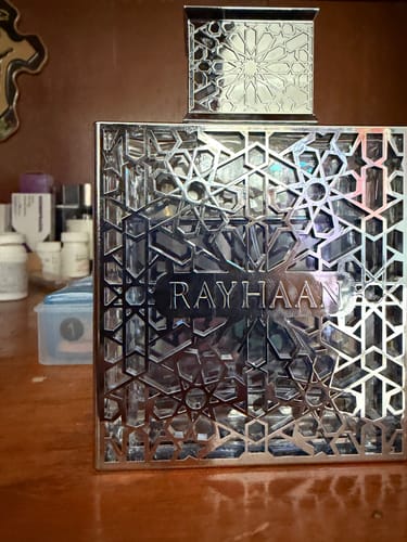Un cliente muestra el frasco del perfume Rayhaan Aquatica con su elaborada cubierta metálica plateada sobre una mesa de madera.