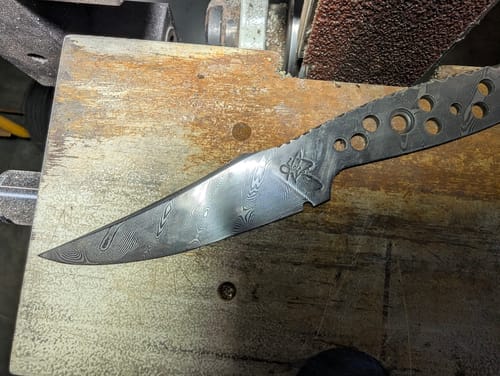 Customer's patterned Damascus knife blade resting in front of a sander with a Flexible Ceramic Sanding Belt.