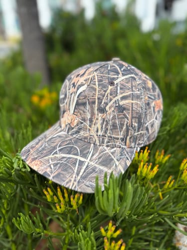 Customer's camouflage-print baseball cap resting on a green plant outdoors.