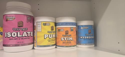 Reviewer's four tubs of Ugly Face supplements, including protein and pre-workout, on a shelf.