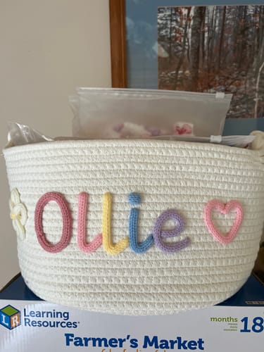 Customer's personalized Baby Knits basket with 'Ollie' in multicolor yarn, flanked by a flower and heart icon.