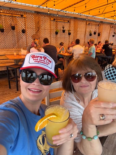 Reviewer wearing the Born Free But Now I'm Expensive trucker hat while enjoying a drink on a sunny patio.