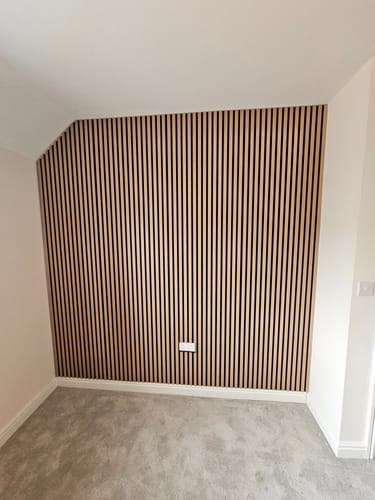 Customer's accent wall covered in light wood Premium Acoustic Slatted Wall Panels, installed in a room with grey carpet.